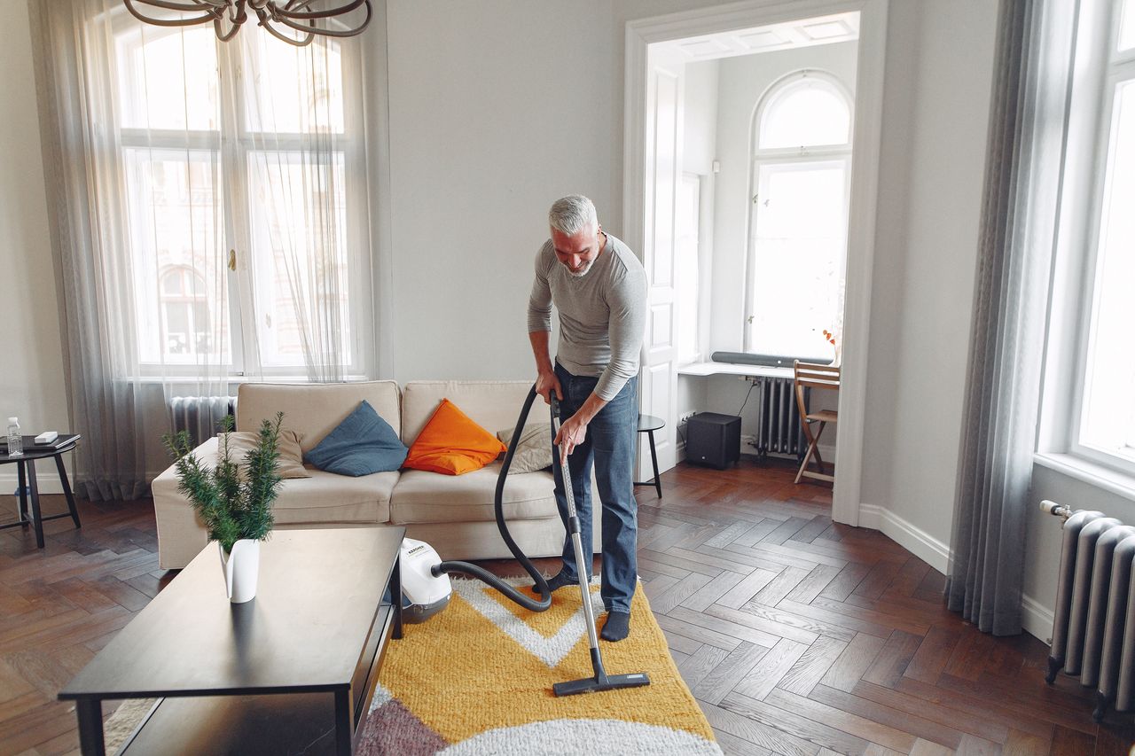 Vue du dessus d’un homme aux cheveux gris et à la barbe, concentré, en train d’aspirer le tapis à la maison